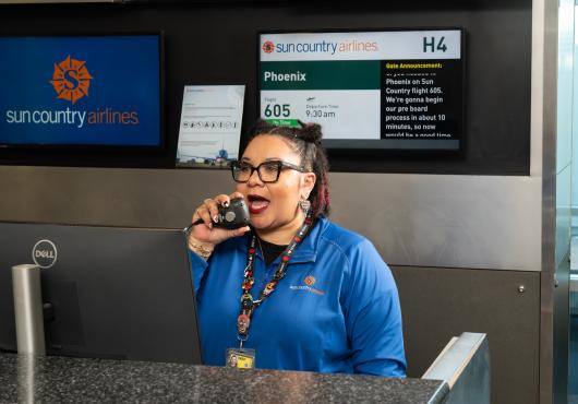 A gate agent is speaking into a handset while her words are displayed on the screen behind her. 